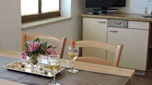 Un comedor con una mesa preparada para dos personas, una bandeja con copas pequeñas y una botella de vino, y un televisor al fondo.