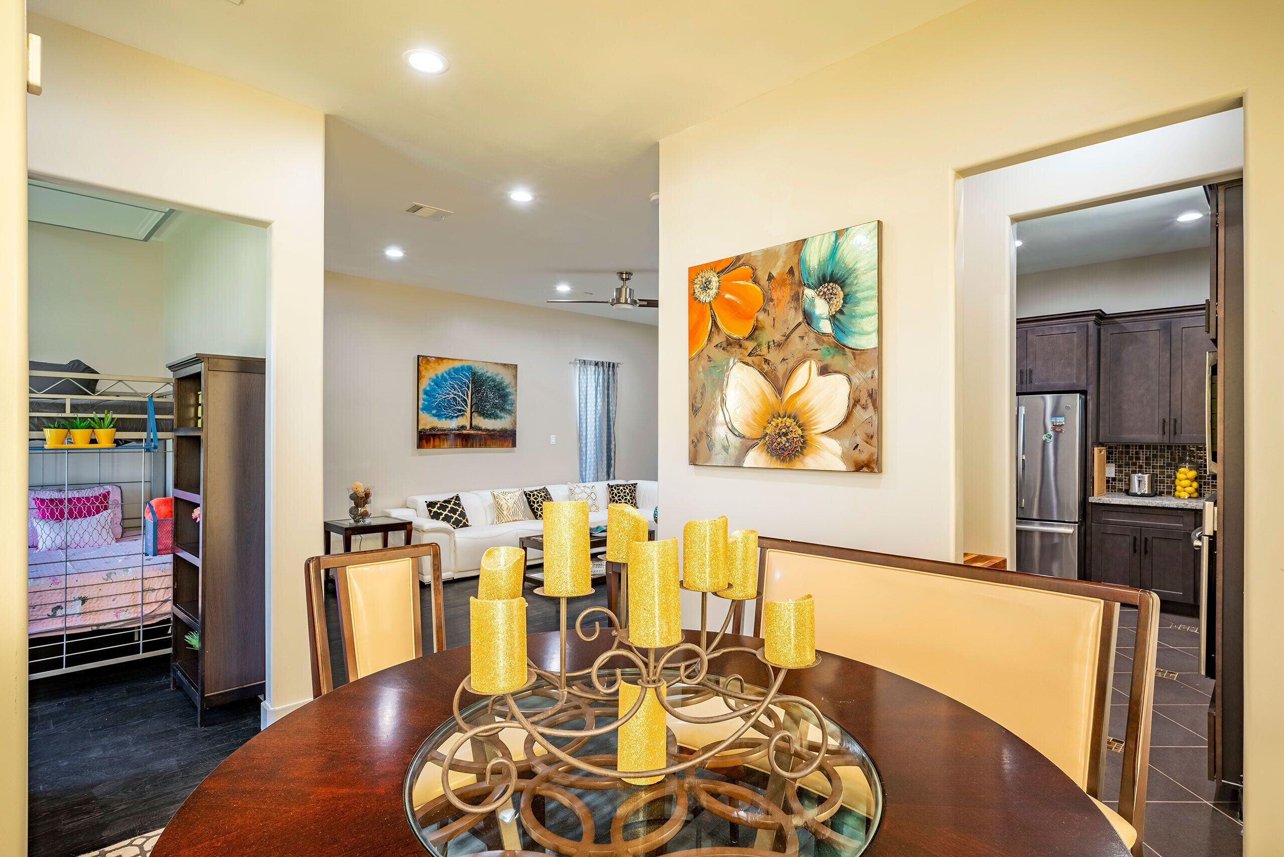 Un comedor con una mesa redonda, sillas y un candelabro decorativo. Hay un cuadro en la pared y una cocina al fondo.