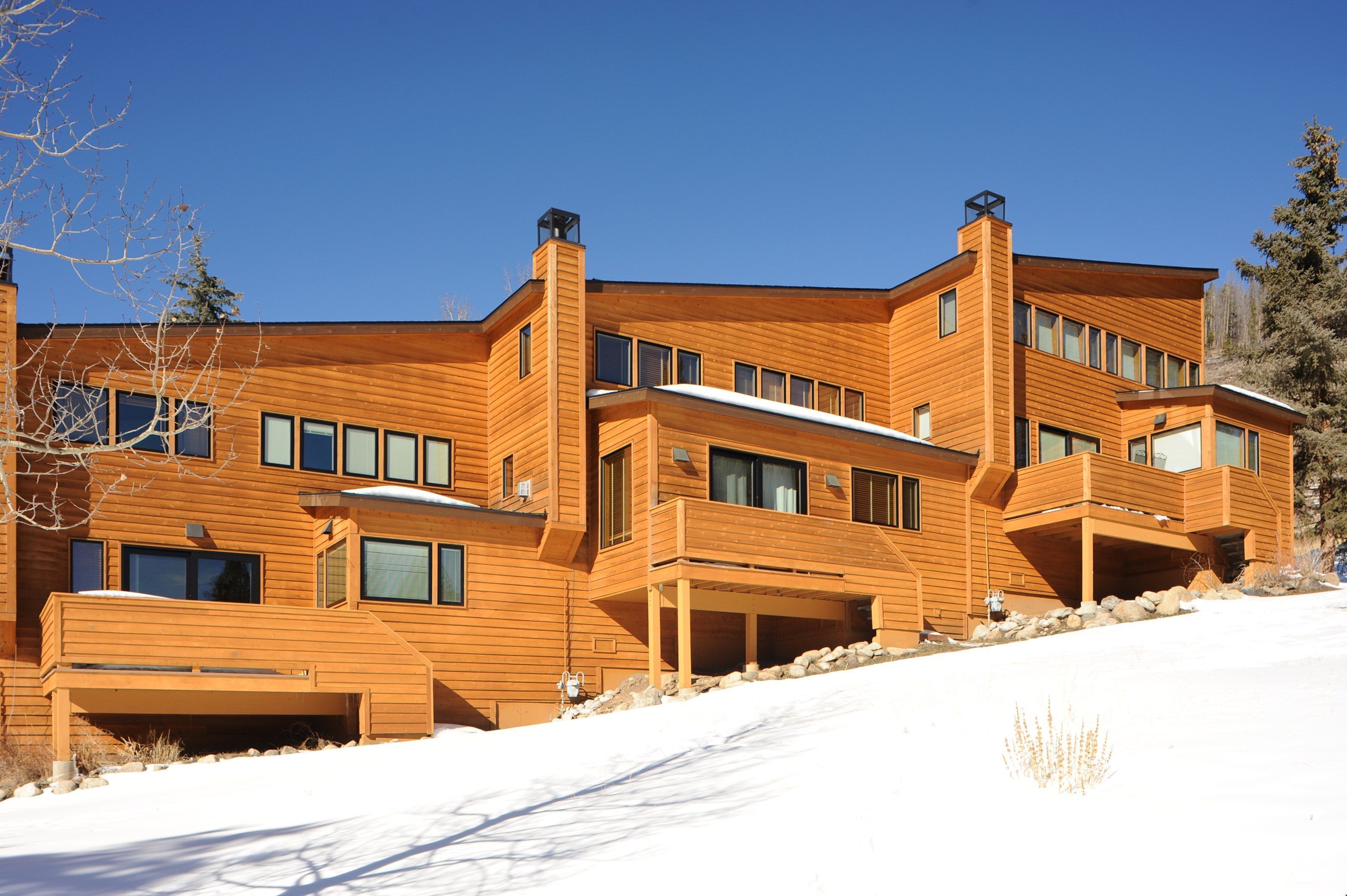 Une maison moderne en bois, à plusieurs niveaux, dotée de grandes fenêtres et d’un espace extérieur couvert.