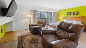 Hotellihuone, jossa on sänky, kaksi nahkaista nojatuolia, pieni pöytä, televisio ja ikkuna, jossa on verhot.