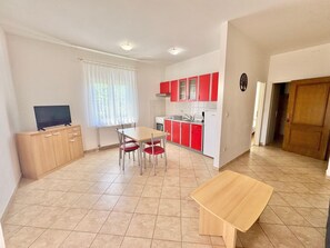 A compact kitchen with red cabinets, a dining table with chairs, a TV on a wooden cabinet, and a wooden bench. - Apartments Cukon (16136-A3) - Medulin (Medulin)