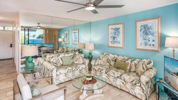 A living room with two patterned sofas, a glass coffee table, a ceiling fan, and framed artwork on the walls.