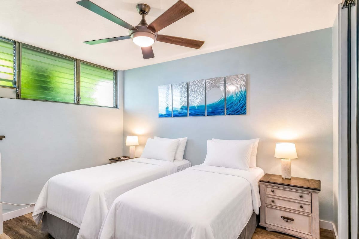 A room with two beds, a ceiling fan, a nightstand with a lamp, and a wall-mounted artwork.