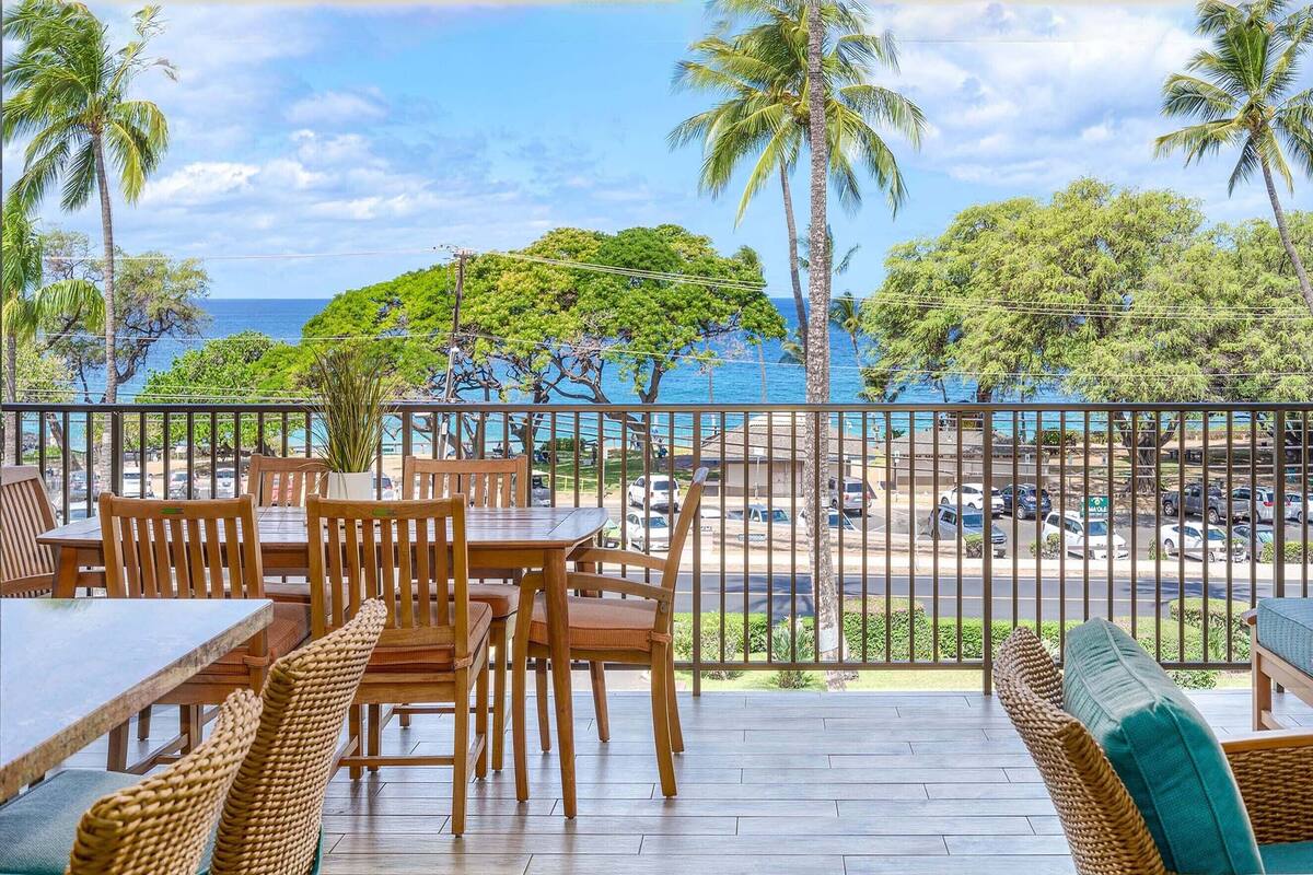 A wooden deck with tables and chairs, overlooking a road and palm trees, with a view of the ocean.
