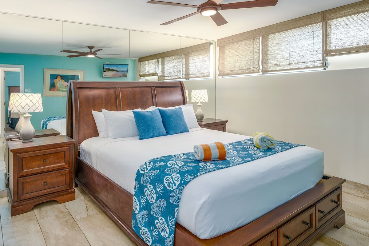 A bedroom with a wooden bed, a ceiling fan, a nightstand with a lamp, and a window with blinds.