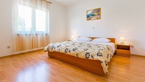 Un dormitorio con una cama de madera, dos mesitas de noche con lámparas, una ventana con cortinas translúcidas y un cuadro en la pared.