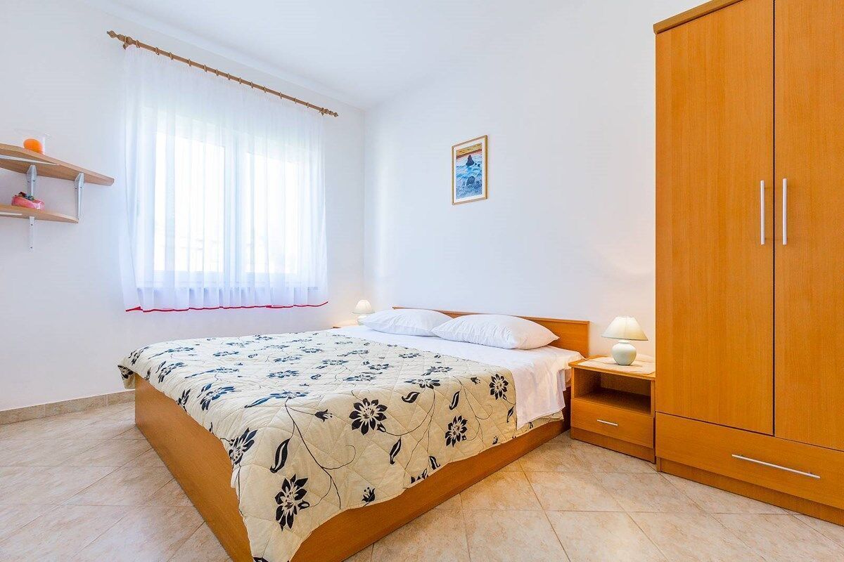 Une chambre à coucher comprenant un lit, une armoire en bois, une table de chevet avec une lampe, une fenêtre avec des rideaux et une étagère fixée au mur.