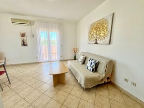 Ein Wohnzimmer mit einer Couch, einem Couchtisch, einem Esstisch und einem großen Fenster mit Vorhängen.