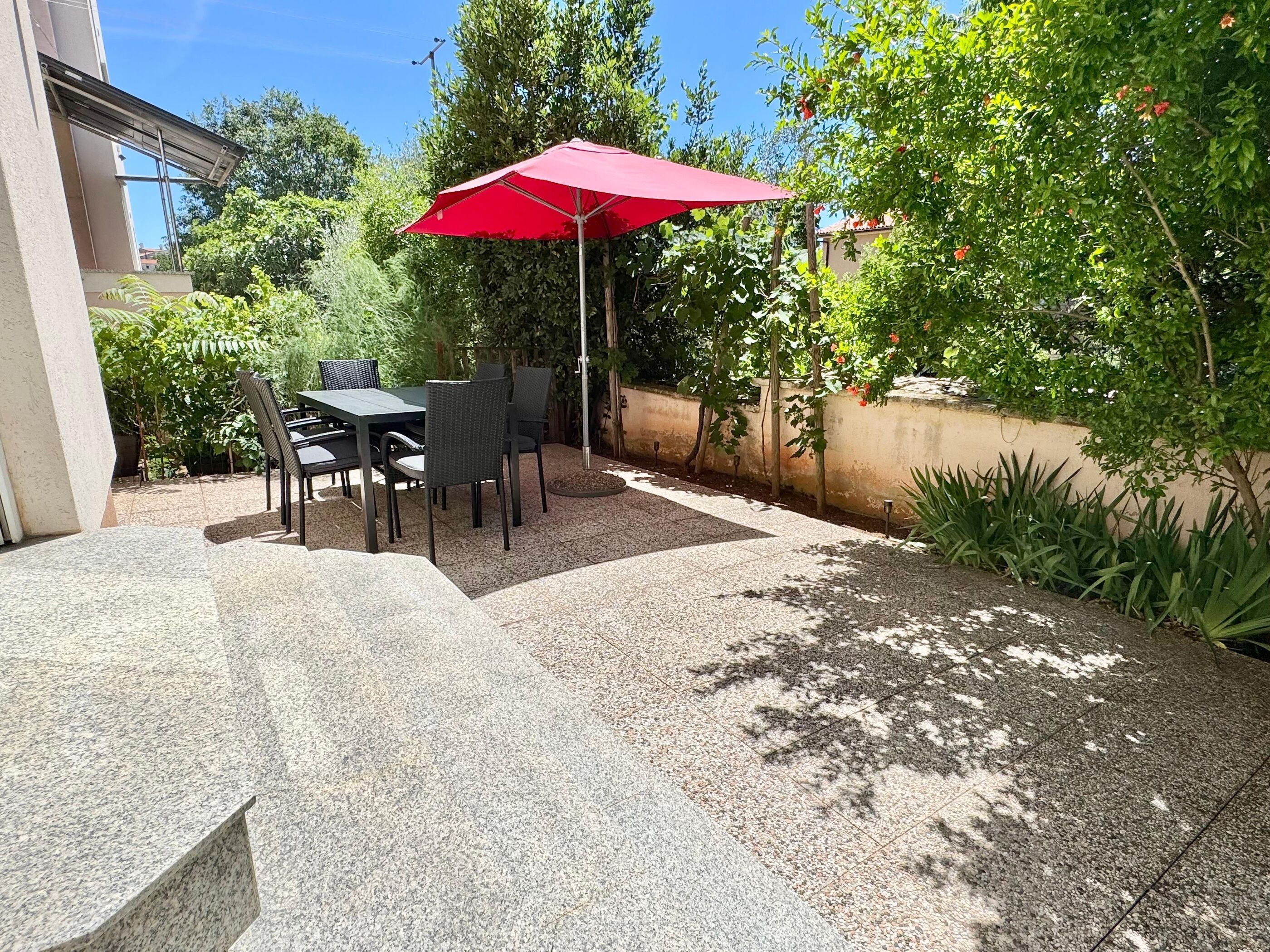 An outdoor patio with a table and chairs, a red umbrella, and surrounding greenery.