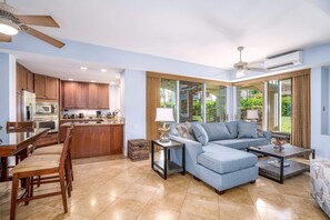 A modern living room with a sectional sofa, a dining table with chairs, and a kitchen area with wooden cabinets and appliances. - The Palms at Wailea (Kihei)