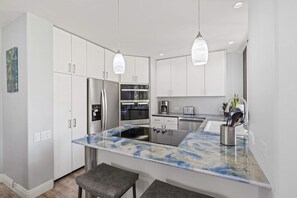 A modern kitchen with white cabinetry, a marble countertop, stainless steel appliances, and pendant lighting. - The Palms at Wailea (Kihei)