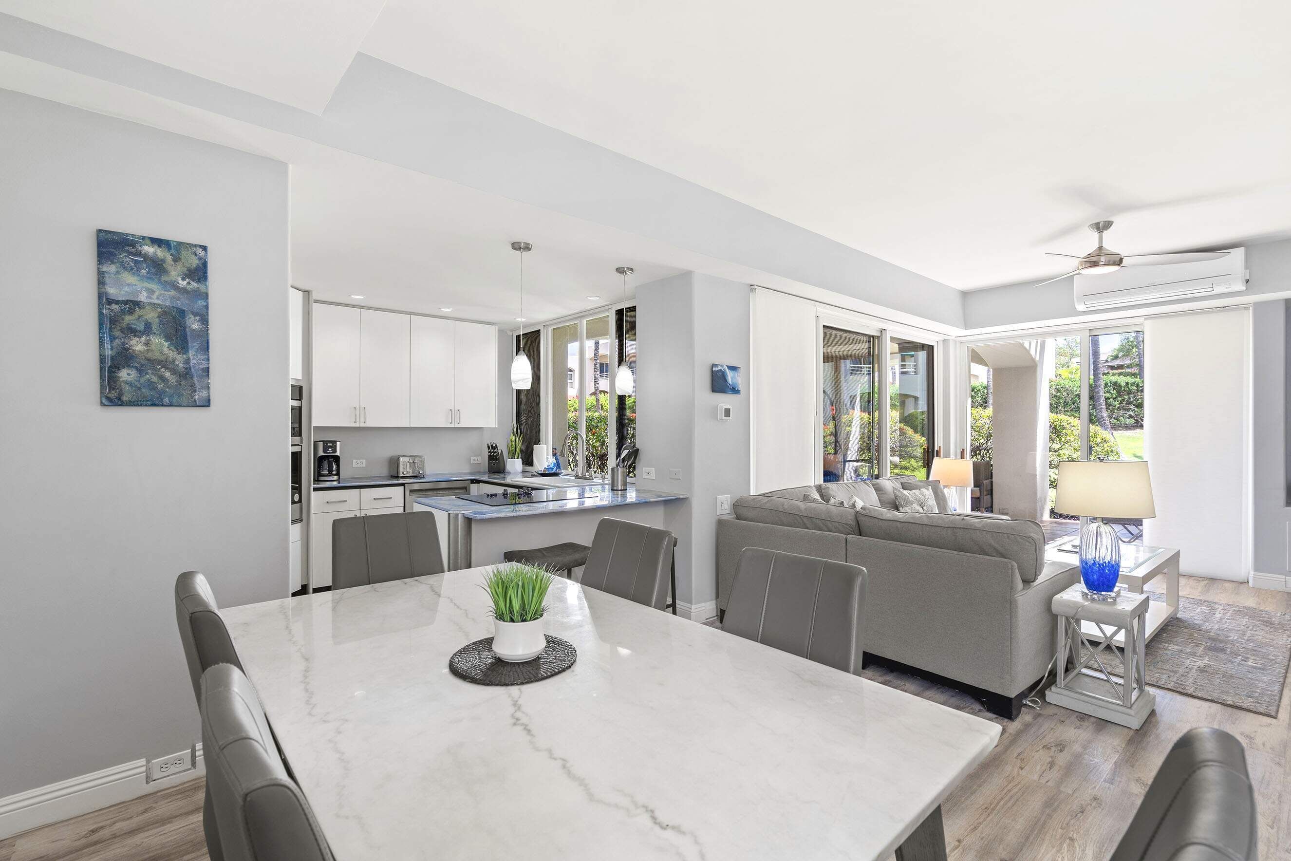 A modern kitchen with white cabinets, a dining table with chairs, and a grey sofa in a bright living area with large windows.