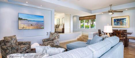 A spacious living room with a large light blue sofa, a ceiling fan, and a dining area with wooden furniture.