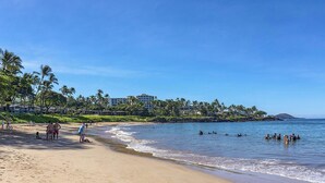 A beach with a hotel in the background, palm trees, and people swimming in the water. - The Palms at Wailea (Kihei)