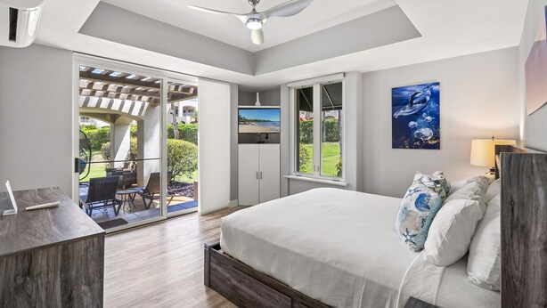 A bedroom with a bed, a desk, a chair, a ceiling fan, and a large window with a view of a garden. - The Palms at Wailea (Kihei)