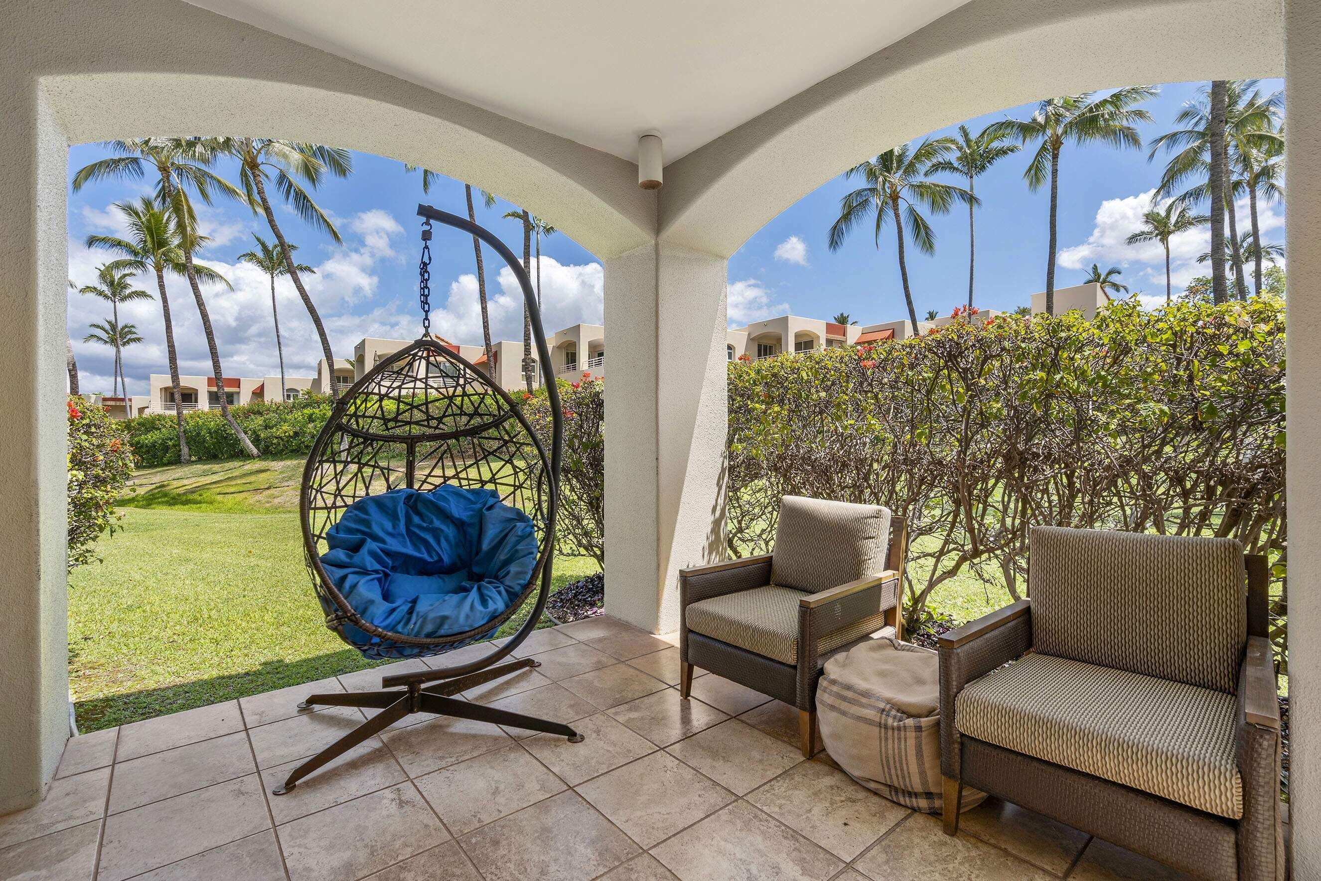 A covered outdoor seating area with a hanging chair, two armchairs, and a view of a landscaped garden with palm trees and a building in the background.