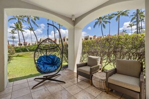 A covered outdoor seating area with a hanging chair, two armchairs, and a view of a landscaped garden with palm trees and a building in the background. - The Palms at Wailea (Kihei)