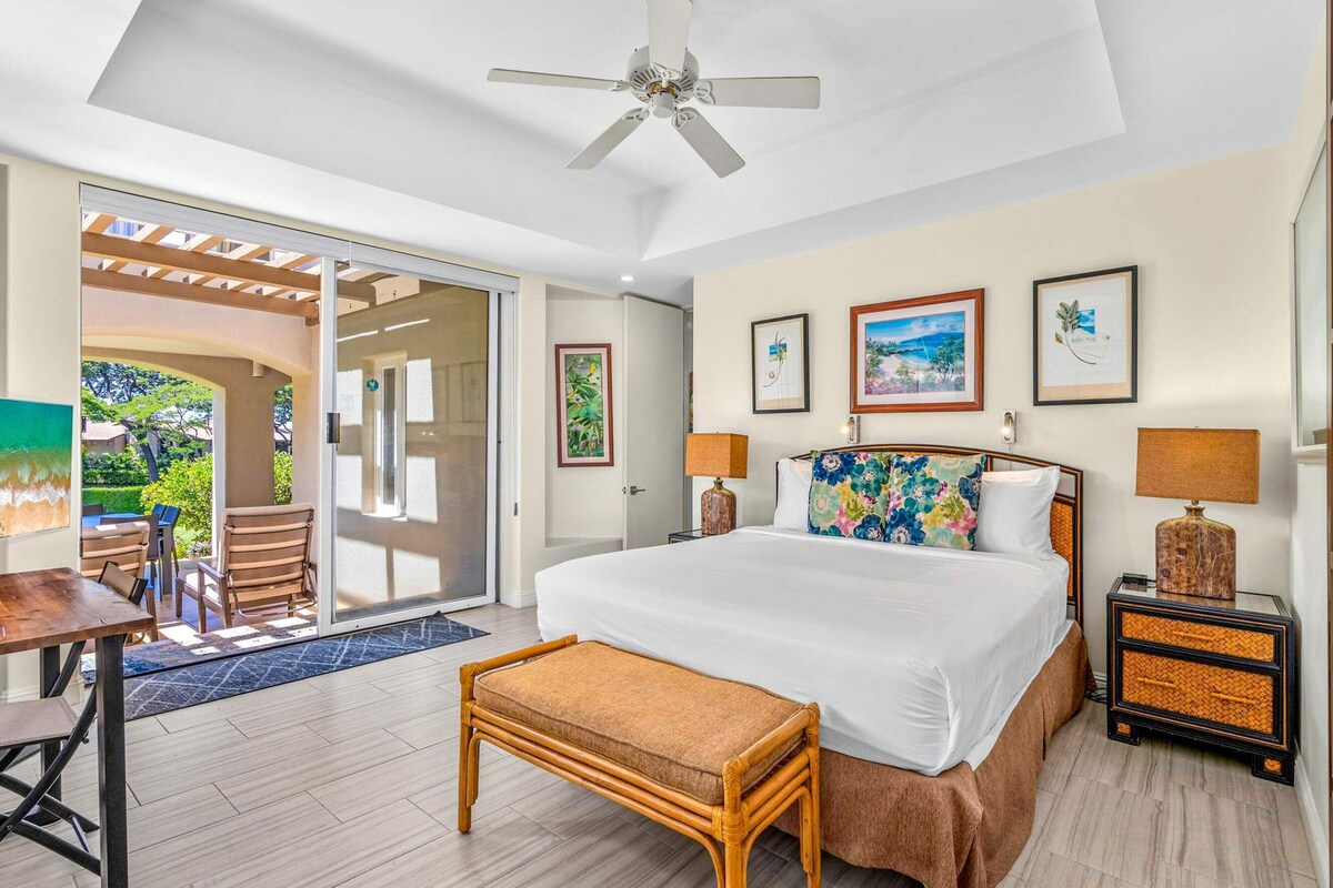 A hotel room with a bed, a desk, a chair, a ceiling fan, and a view of the outdoors.
