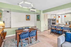 A modern kitchen with built-in appliances, a dining area with a wooden table and blue chairs, and a living area with a sofa and a coffee table. - The Palms at Wailea (Kihei)