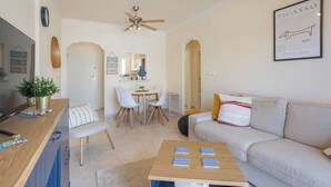 A modern living room with a sofa, a coffee table with tiles, a dining area with chairs and a table, and a kitchen area with a counter and cabinets. - cozy Ground-floor apartment 4p terrace pool & parking beach --> 2km (VC)