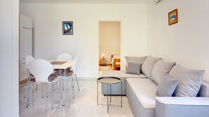 Ein modernes Wohnzimmer mit grauem Sofa, weißen Sesseln, einem kleinen runden Tisch und einem Essbereich mit Tisch und Stühlen. An der Wand hängt ein Gemälde und unter der Decke befindet sich eine Klimaanlage.