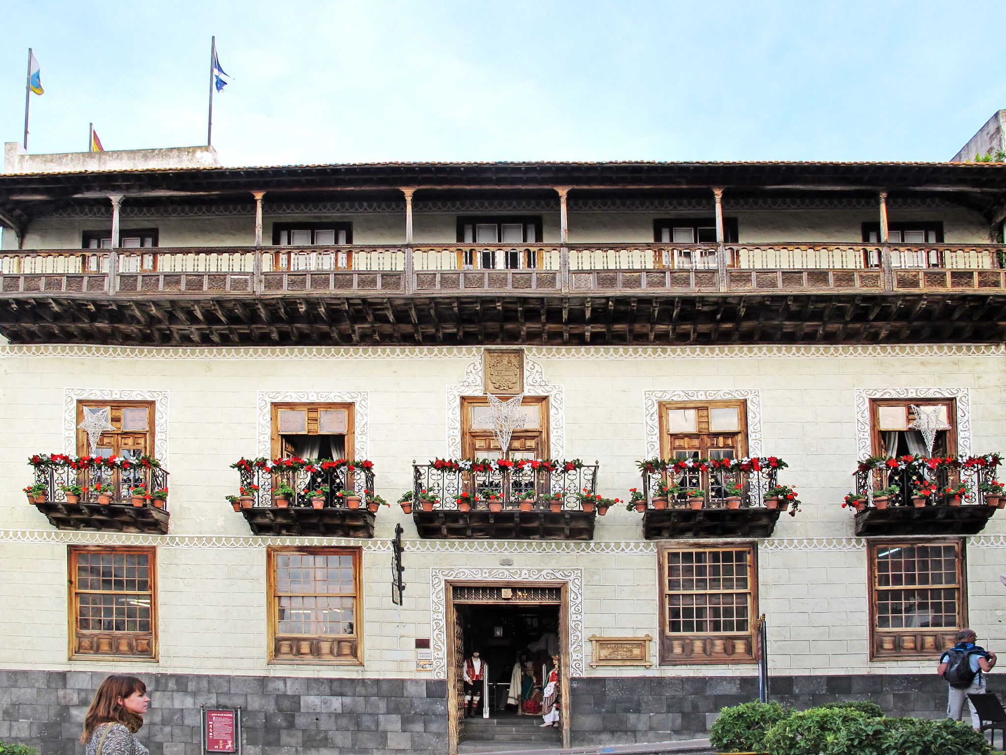 A traditional building with multiple balconies adorned with red flowers, wooden doors, and a stone facade.