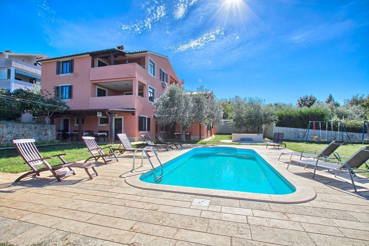 A swimming pool with lounge chairs, a pink building, and a clear blue sky.