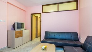 Ein kleines Wohnzimmer mit einer Couch, einem Couchtisch mit Blumen, einem Fernseher auf einem Schrank und einer Tür zu einem weiteren Raum.