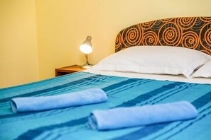Un lit bien fait, avec une literie bleue, un oreiller blanc et une tête de lit ornée d’un motif en spirale.