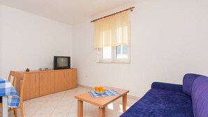 Ein kleiner Wohnraum mit einer Couch, einem Fernseher auf einem Holzschrank, einem Tisch mit Obstschale und einem Fenster mit Vorhängen.