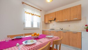 A compact kitchen with wooden cabinets, a dining table with a pink tablecloth, and a window with a curtain. - Apartments Verde (16211-A2) - Premantura (Premantura)