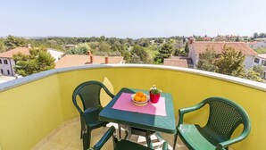 Un balcon avec un mur jaune, deux chaises vertes, une table avec un saladier de fruits et une plante en pot, et une vue sur un quartier résidentiel.