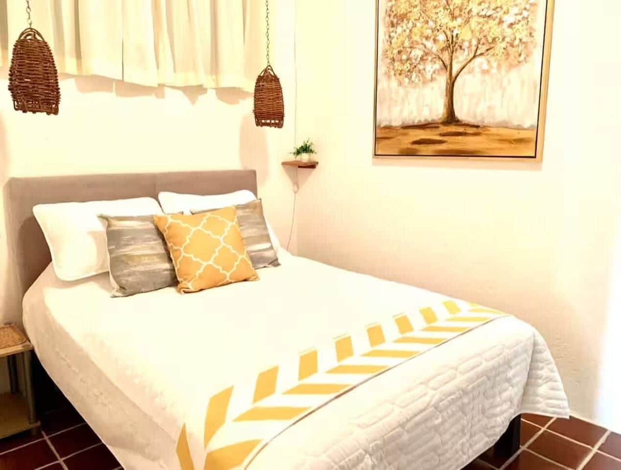 Una cama con cubrecama blanco y almohadas decorativas, una cesta colgante, una planta pequeña y un cuadro en la pared.