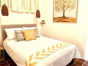 A bed with a white bedspread and decorative pillows, a hanging basket, a small plant, and a framed picture on the wall. - Villas Mimosa (Zihuatanejo)