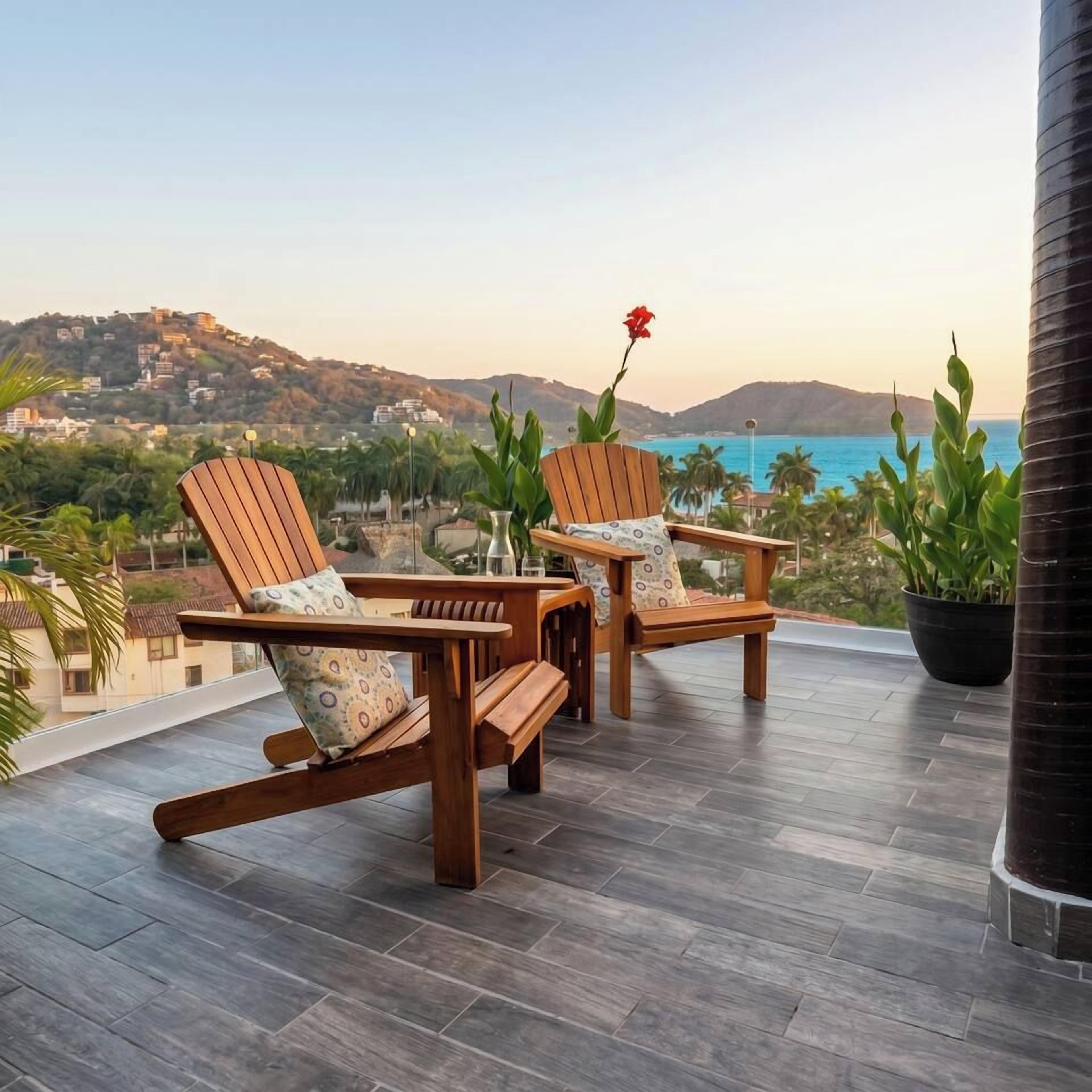 A terrace with two wooden lounge chairs, a potted plant, and a view of the sea and hills.