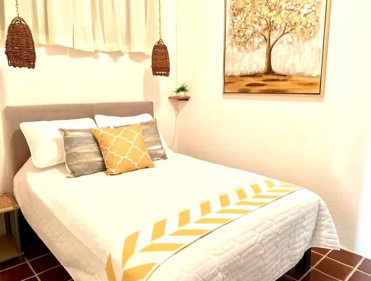 A bed with a white bedspread and decorative pillows, a hanging basket, a small plant, and a framed picture on the wall.