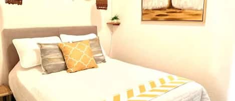 A bed with a white bedspread and decorative pillows, a hanging basket, a small plant, and a framed picture on the wall.