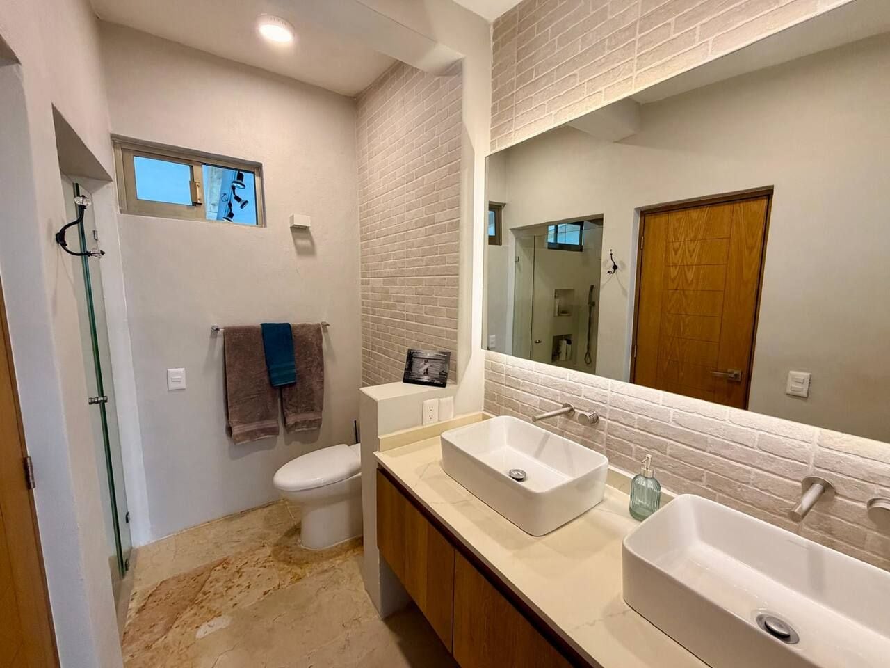 A modern bathroom with a large mirror, two sinks, a toilet, and a shower area.