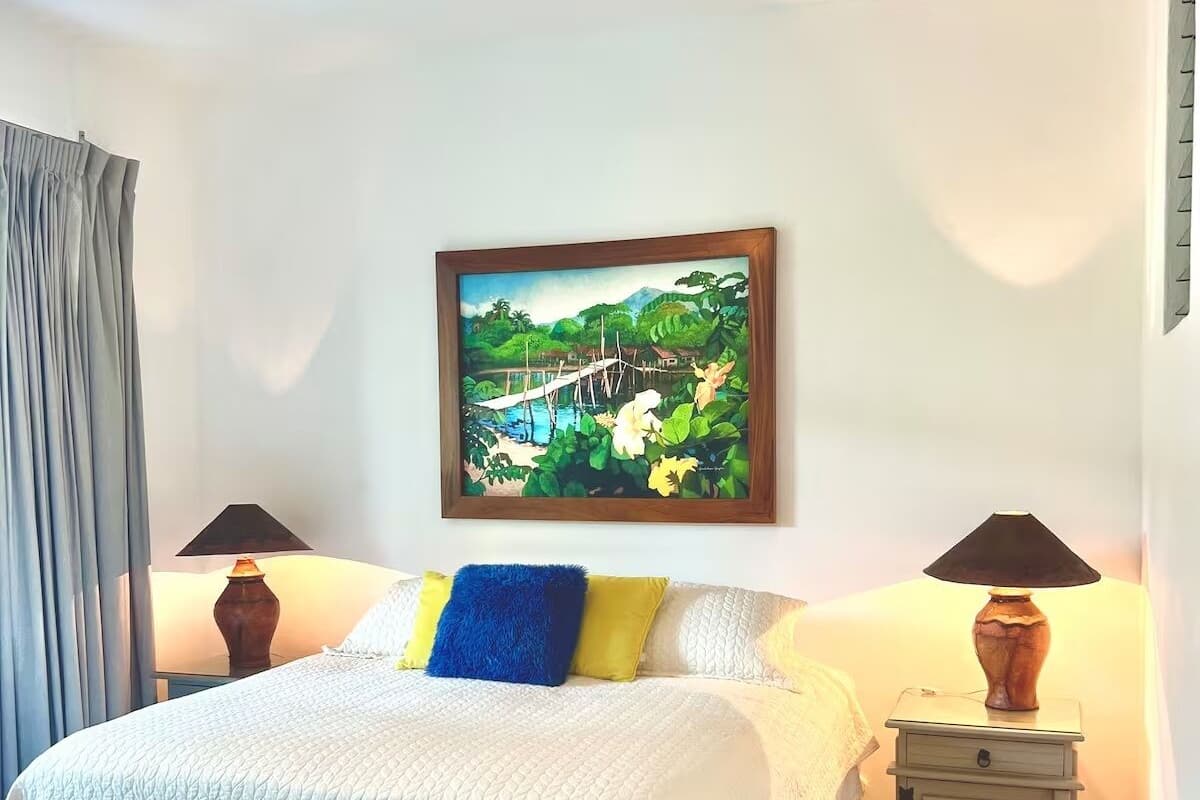 Een slaapkamer met een bed, een plafondventilator, een schilderij, twee nachtkastjes met lampen en een raam met gordijnen.