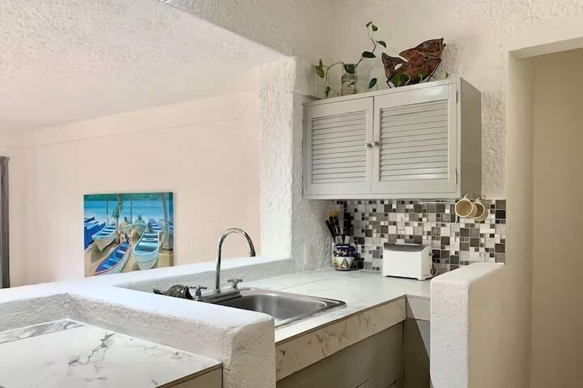 Een keuken met witte kasten, een betegelde spatwand en een schilderij van boten aan de muur.