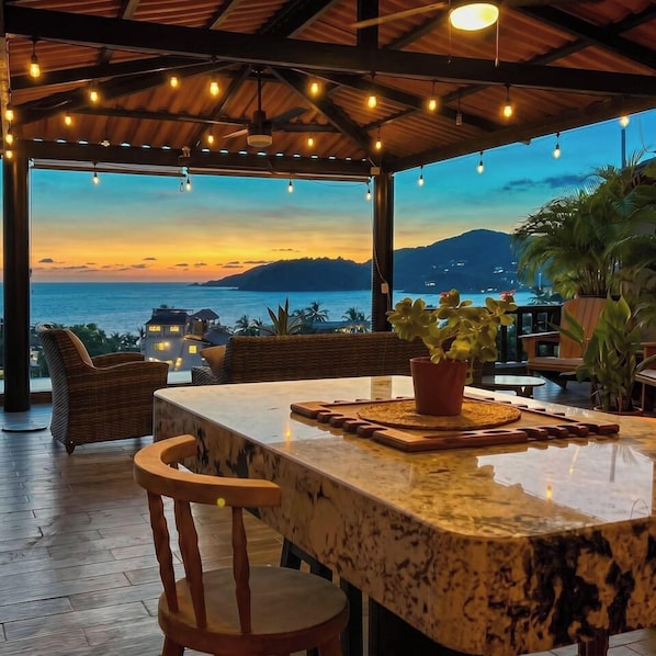 Un área de estar al aire libre con vistas al mar y a las montañas, que incluye una mesa y sillas de madera.