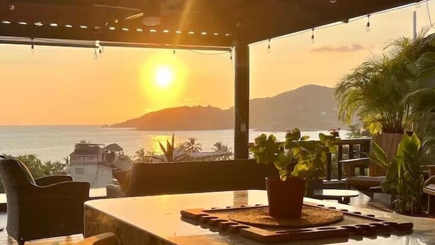 Una terraza iluminada por el sol con vistas al mar y a las montañas, que incluye una mesa de mármol, sillas y plantas en macetas.