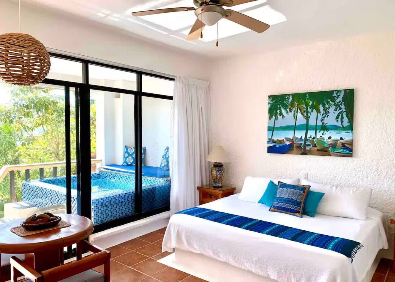 A hotel room with a bed, a table, a chair, a balcony with a pool, and a painting of a beach scene.
