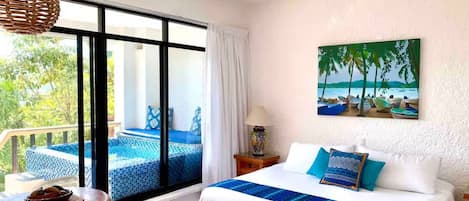 A hotel room with a bed, a table, a chair, a balcony with a pool, and a painting of a beach scene.