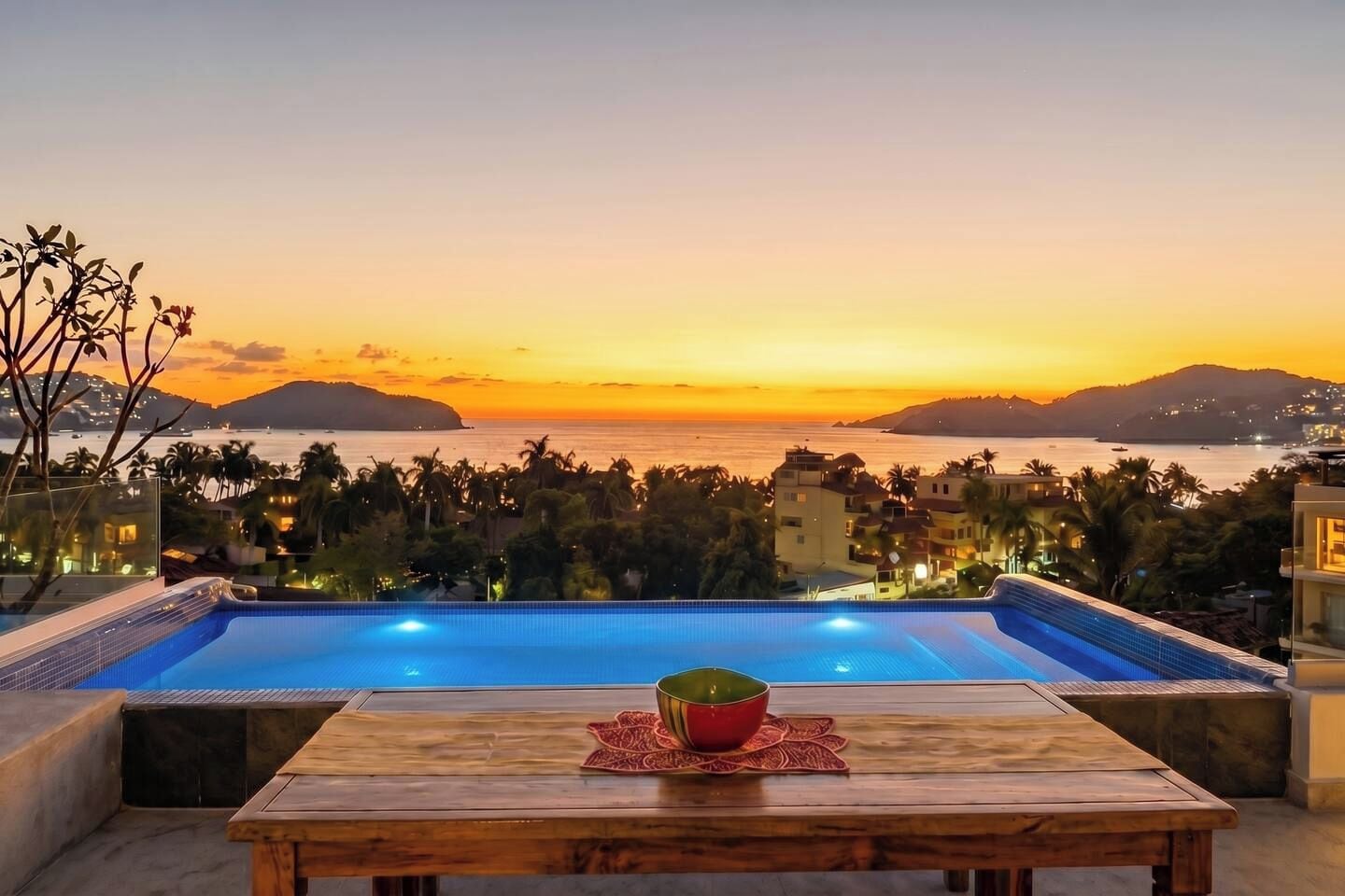 An infinity pool with a wooden table and a bowl, overlooking a coastal cityscape at sunset.