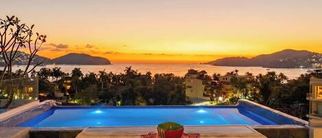 An infinity pool with a wooden table and a bowl, overlooking a coastal cityscape at sunset.