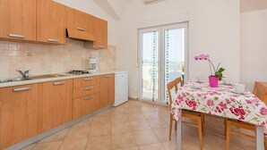 A modern kitchen with wooden cabinets, a gas stove, and a small refrigerator. There is a dining area with a table set for two, a pink flower vase, and a view through a glass door. - Apartments Verde (16211-A5) - Premantura (Premantura)