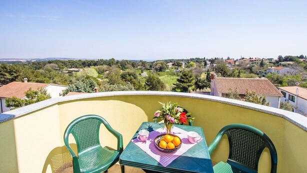 A balcony with two green chairs, a table with a bowl of fruit and a vase of flowers, and a clear view of a residential area. - Apartments Verde (16211-A5) - Premantura (Premantura)