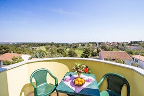 Un balcó amb dues cadires verdes, una taula amb un plat de fruita i un vas de flors, i una vista despejada d'una zona residencial.
