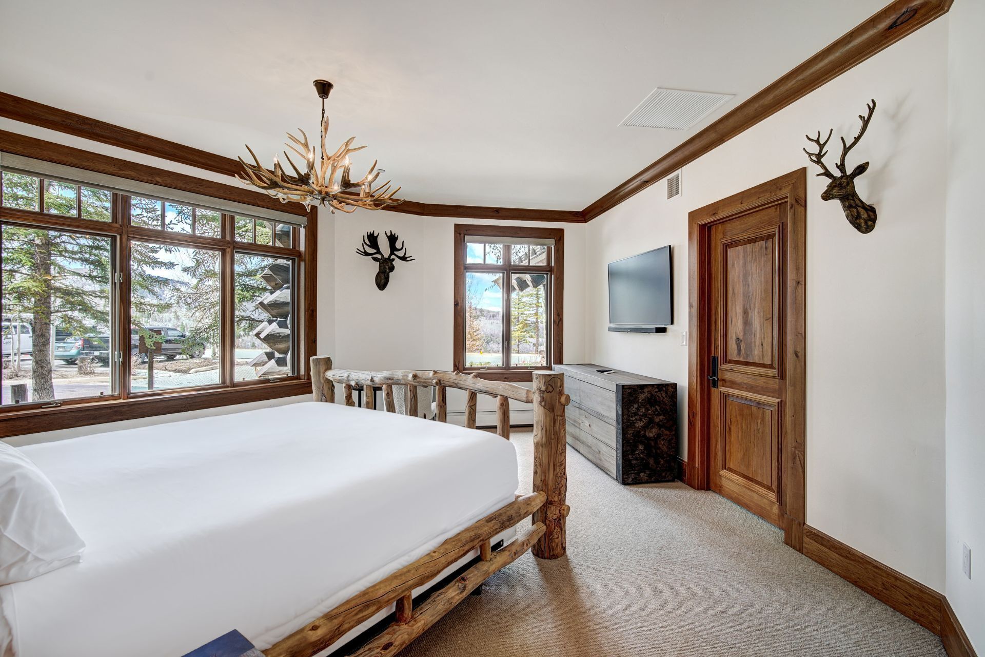 A bedroom with a large bed, wooden headboard, a chandelier, a desk with a chair, a television, and deer antlers on the wall.
